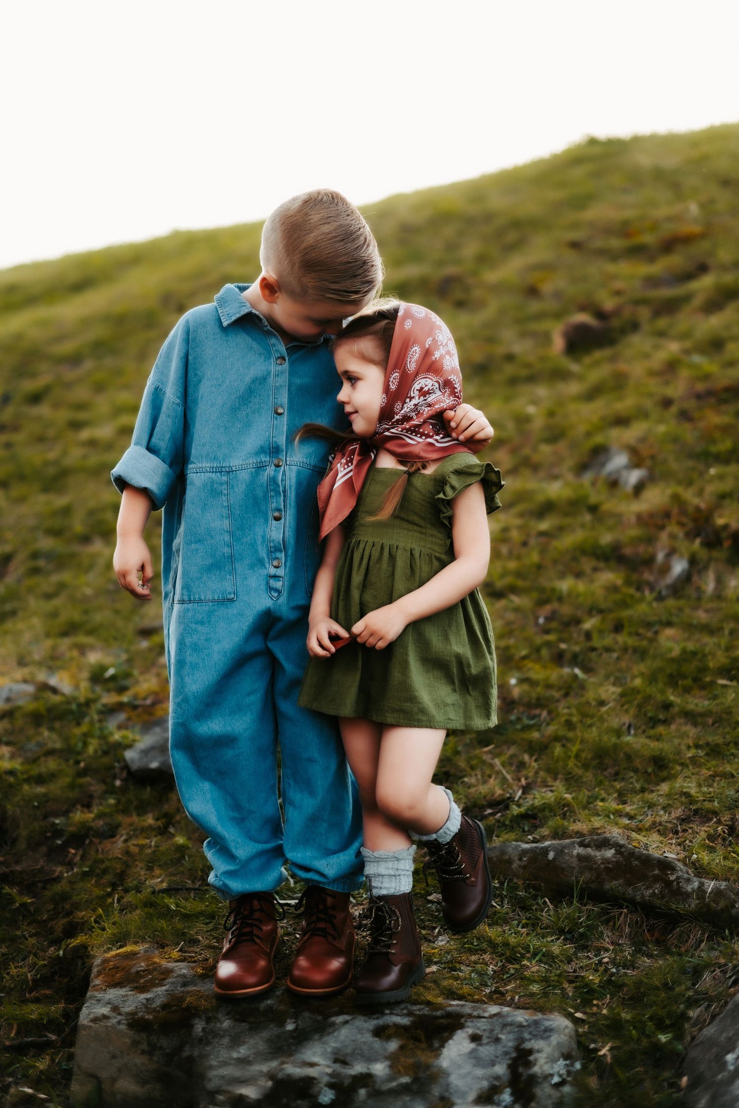 Two children in vintage-inspired outfits stand together on a mossy hill with their arms around each other.