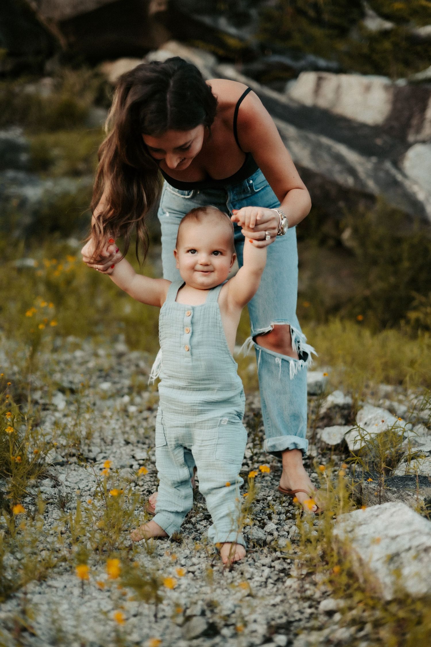 Parent and baby in matching light denim outfits spending time together outdoors among rocks and wildflowers.