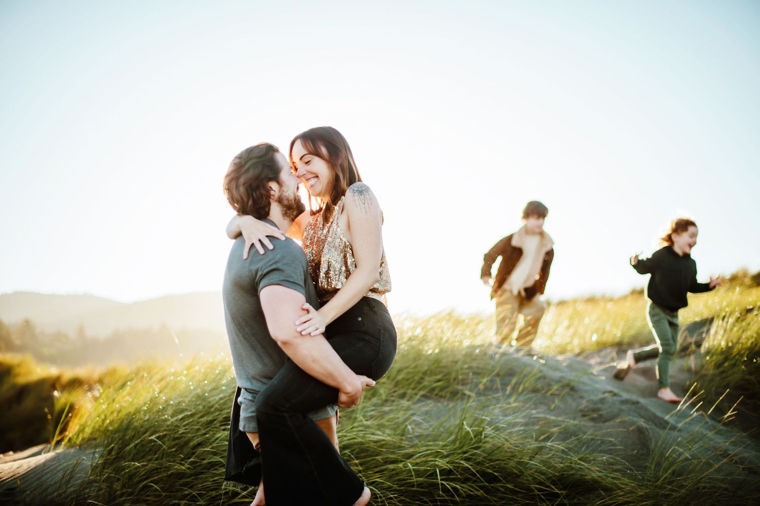 Happy couple embraces on grassy hilltop at sunset while friends walk in background during golden hour.