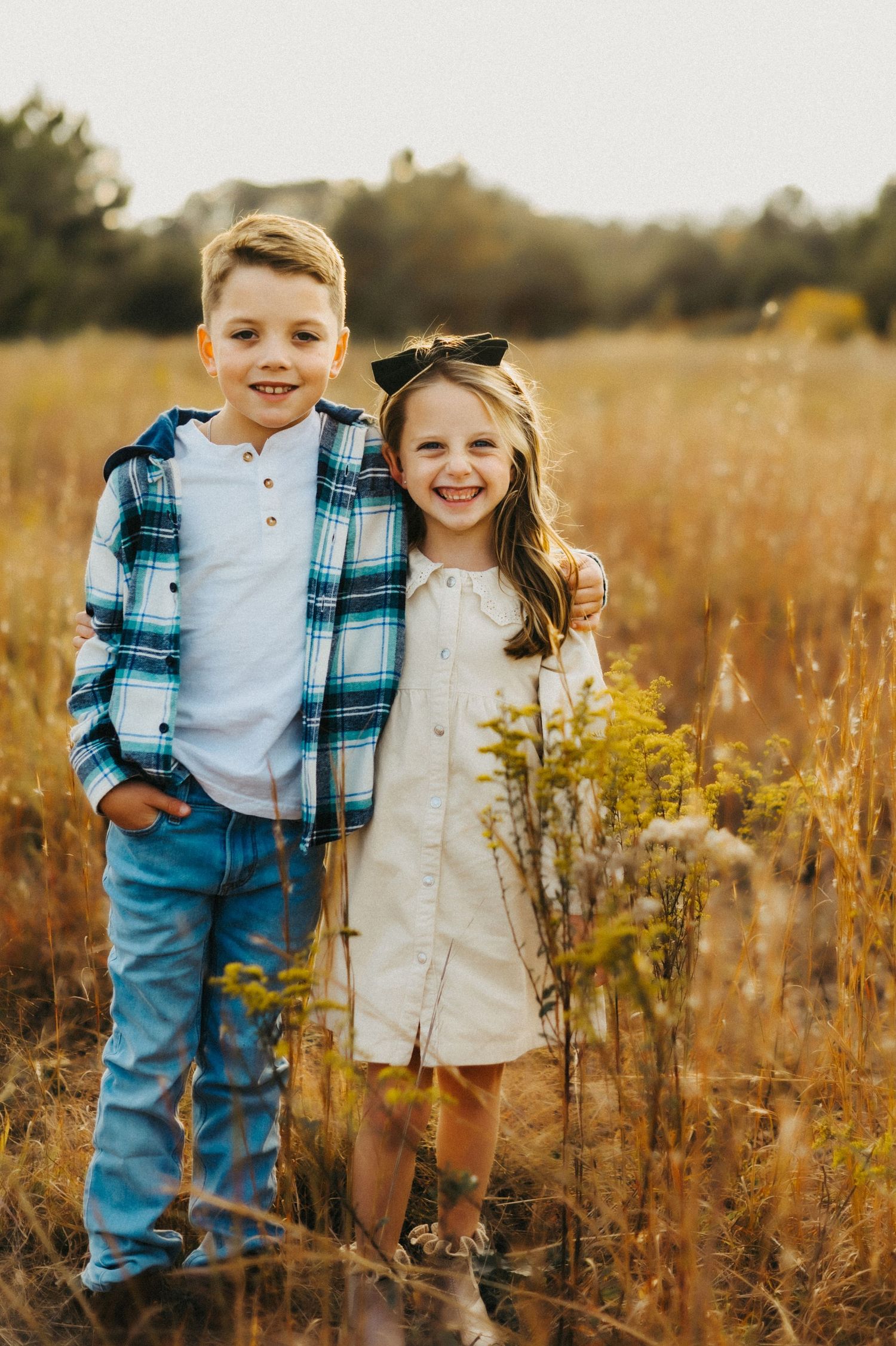 Two children stand together in tall golden grass at sunset wearing casual clothes on an autumn evening.