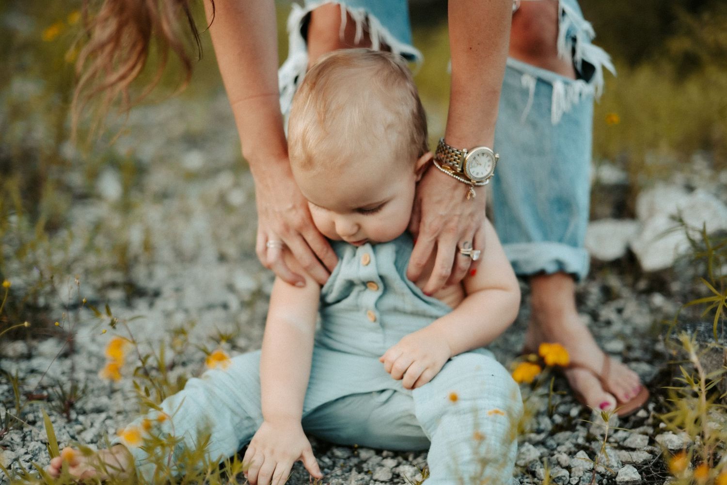 Baby in light blue overalls sits on the ground exploring yellow wildflowers while parent holds them securely.