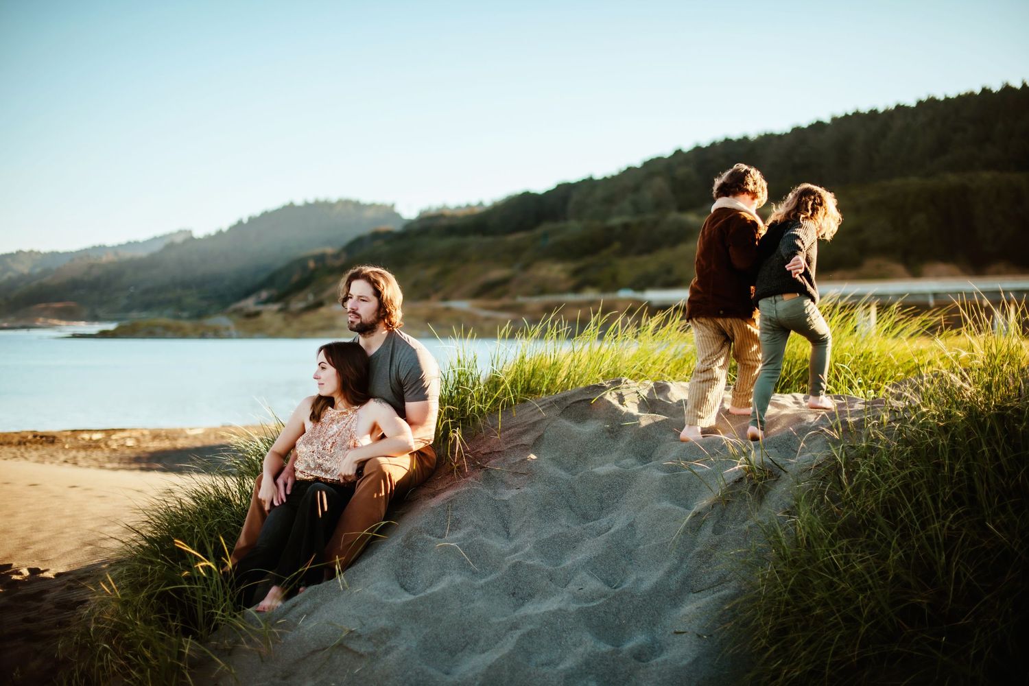 People enjoying a warm sunset on sandy coastal dunes with ocean and mountains in background.