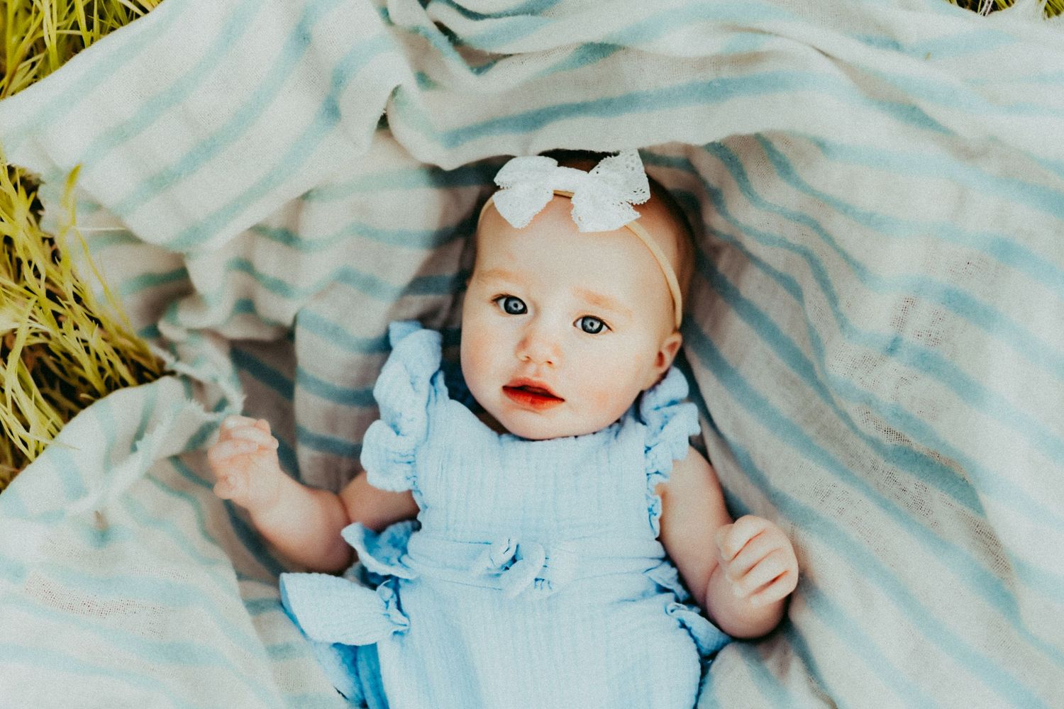 A baby in a light blue dress lies on a striped blanket with a white bow hair accessory.