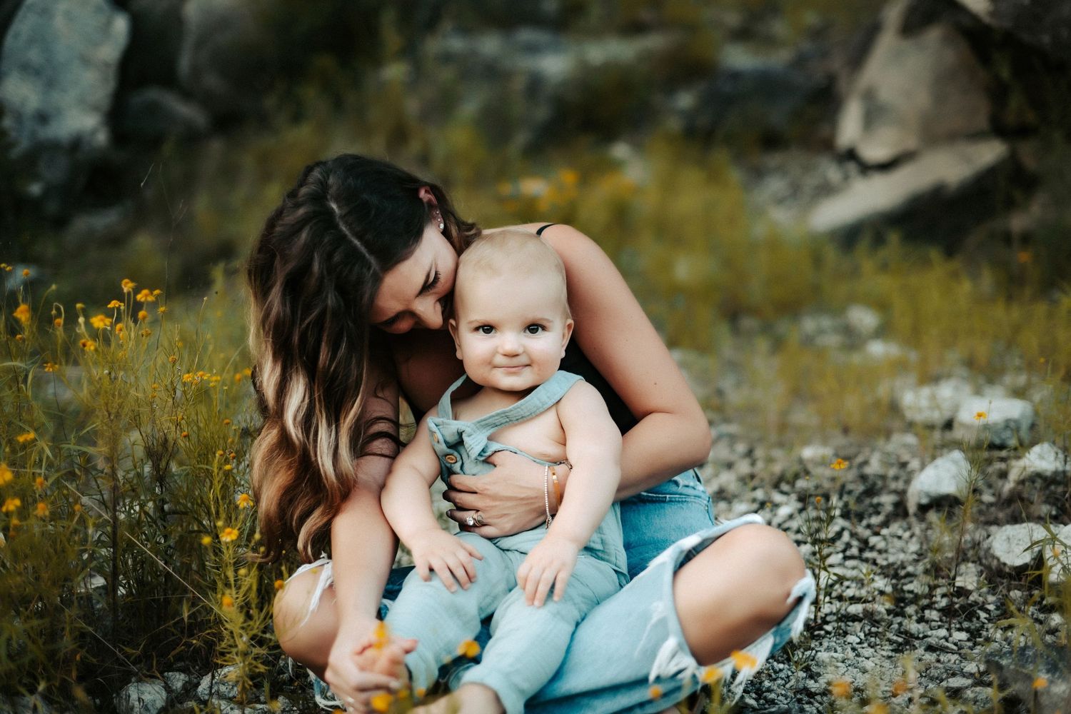 A mother and baby sit in a meadow surrounded by wildflowers on a sunny afternoon.