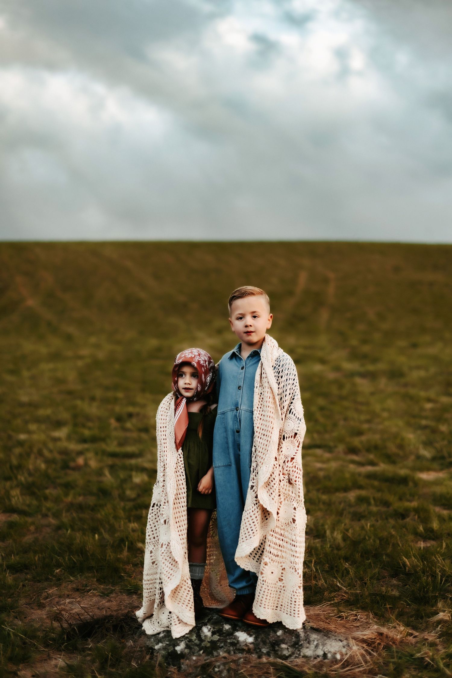 Two children stand together wrapped in a cream knitted blanket on a grassy hilltop under cloudy skies.