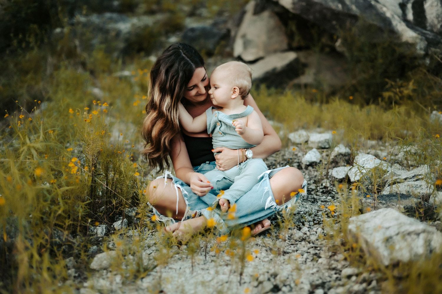 Two people share a tender moment while sitting among yellow wildflowers and rocks in a natural outdoor setting.