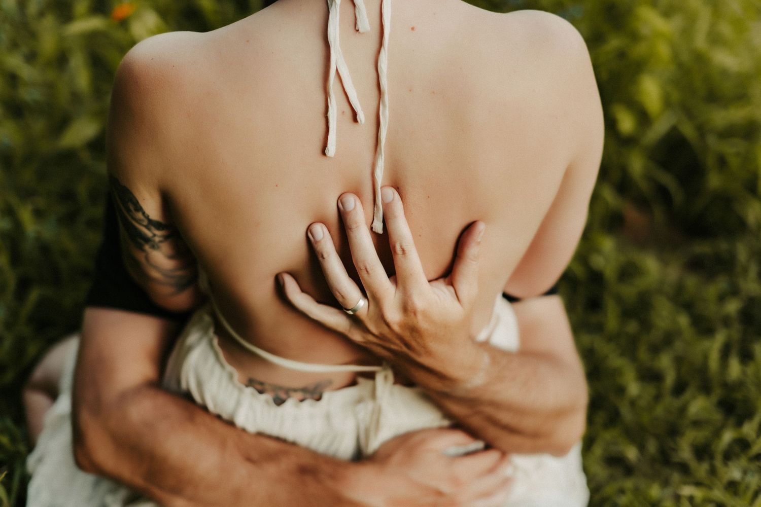 Intimate embrace captured from behind showing bare shoulders and hands against green foliage background.