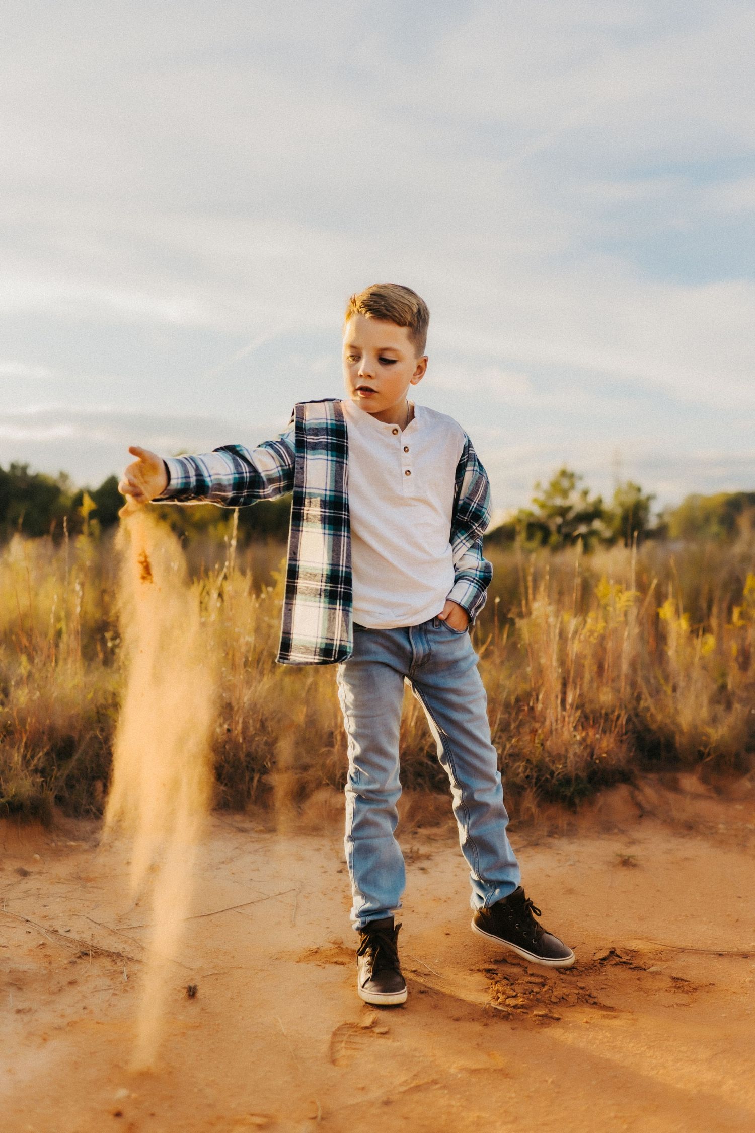 Playing in soft sand at sunset, a child in jeans and plaid shirt tosses dust into golden sunlight on a warm evening.