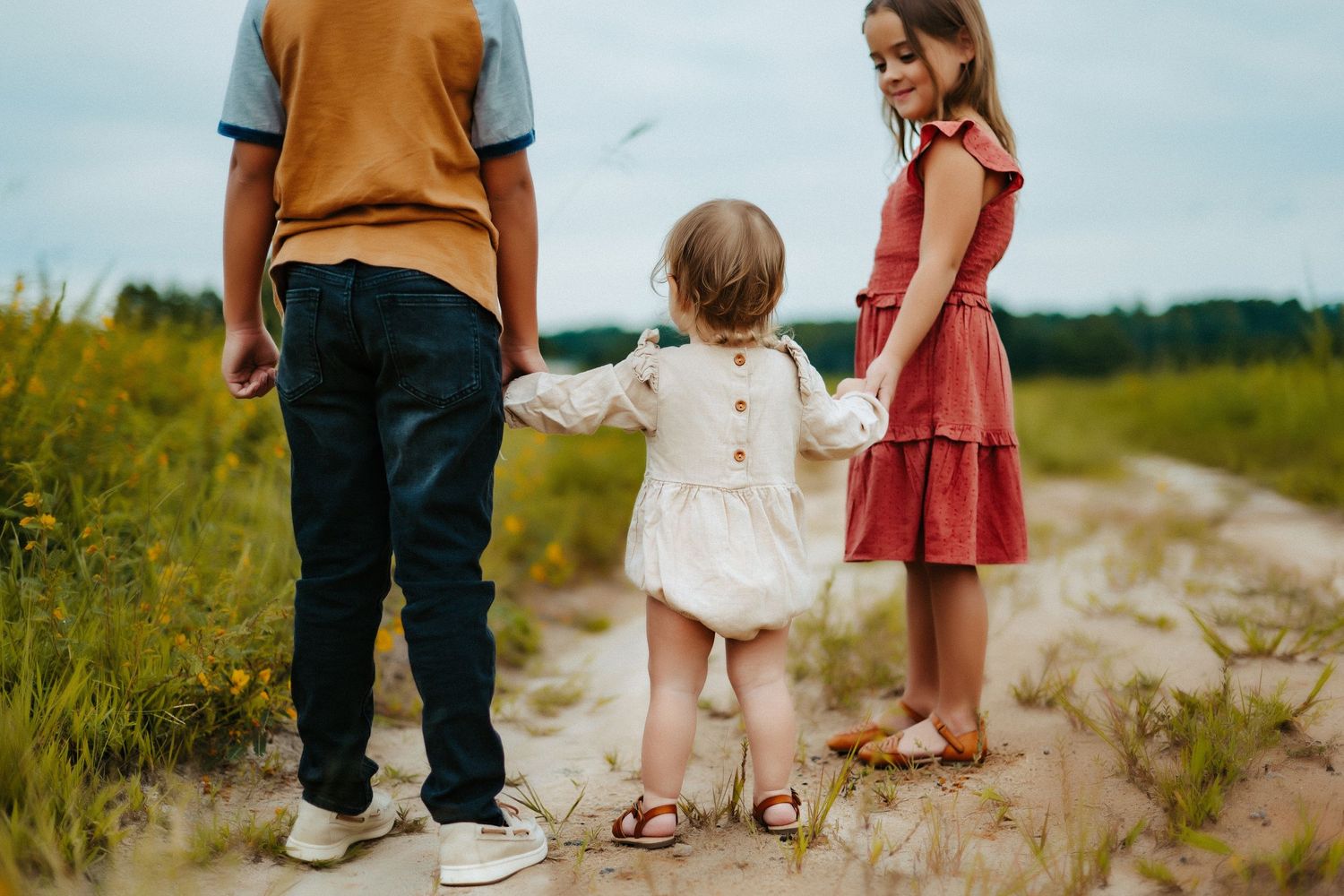 Three people walk together on a dirt path through a grassy meadow on a summer day.