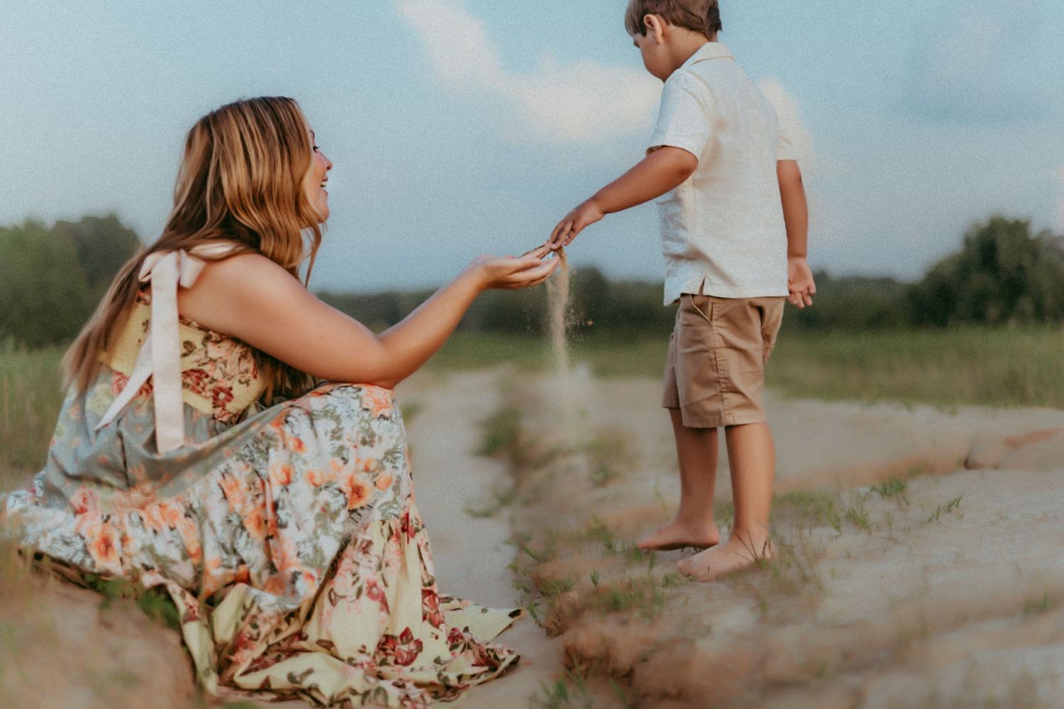 Two people share a tender moment while standing in a dusty field at sunset, one wearing a floral dress and another in shorts.