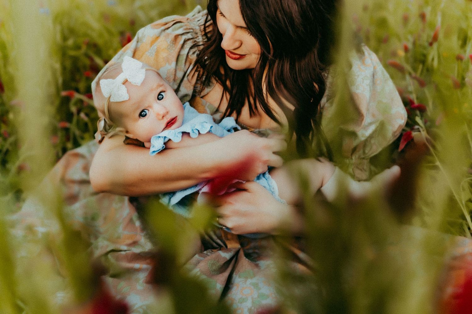 Mother and baby smiling together outdoors surrounded by green foliage on a sunny day.