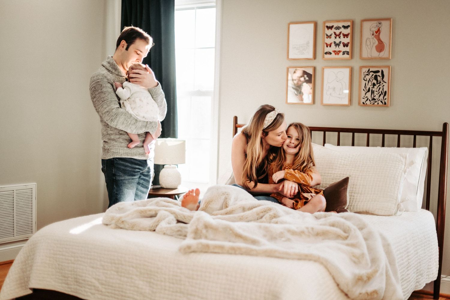 A family enjoying a relaxed morning together in a cozy bedroom with framed wall art and white bedding.