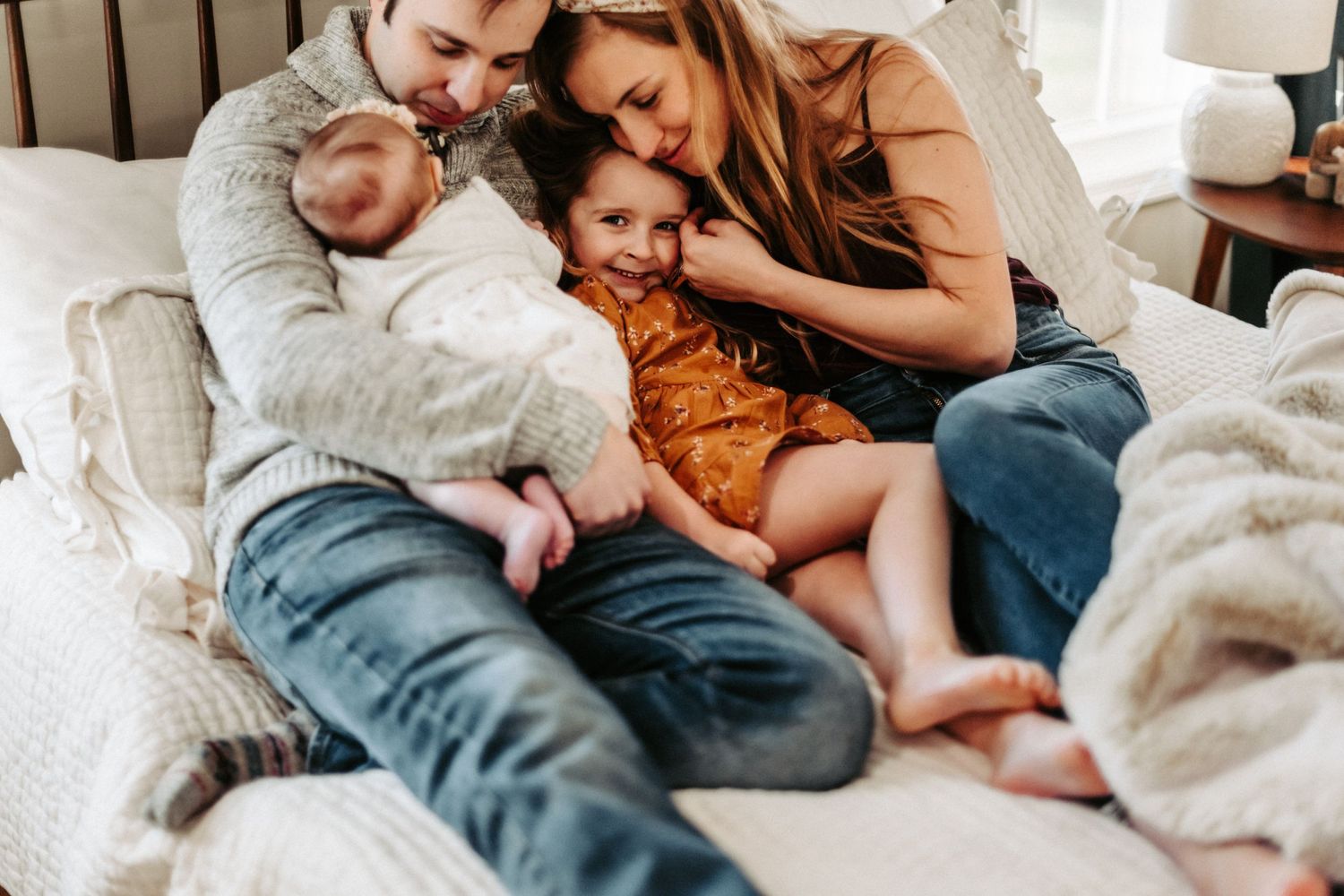 Cozy family cuddles together on white bed during lifestyle photoshoot.