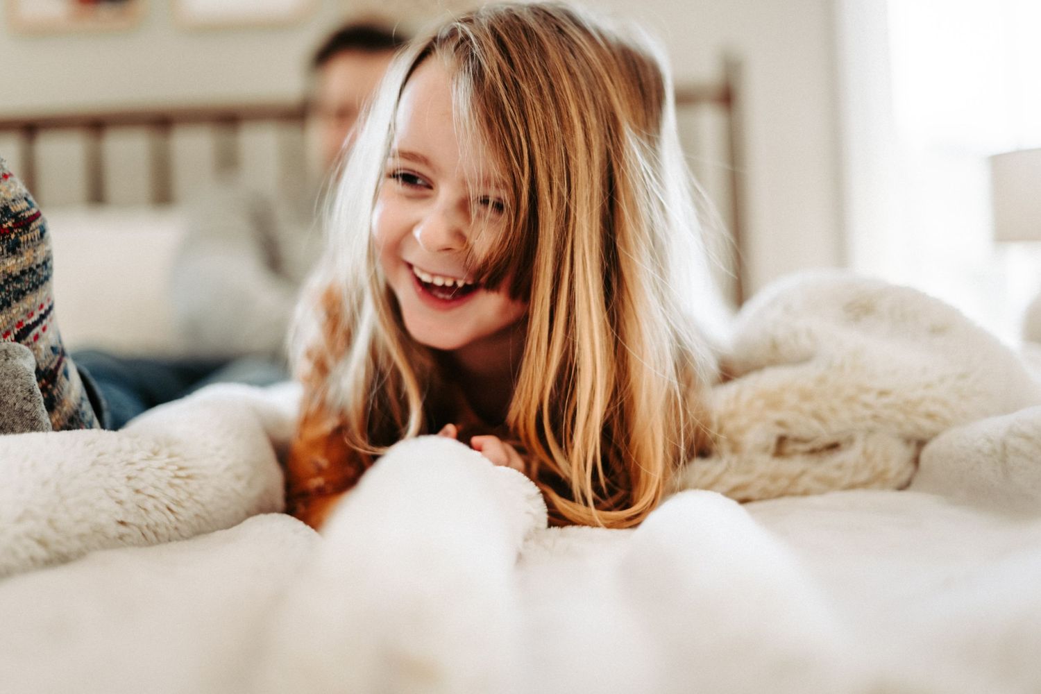 A cheerful person lying on white bedding smiles brightly while relaxing at home.