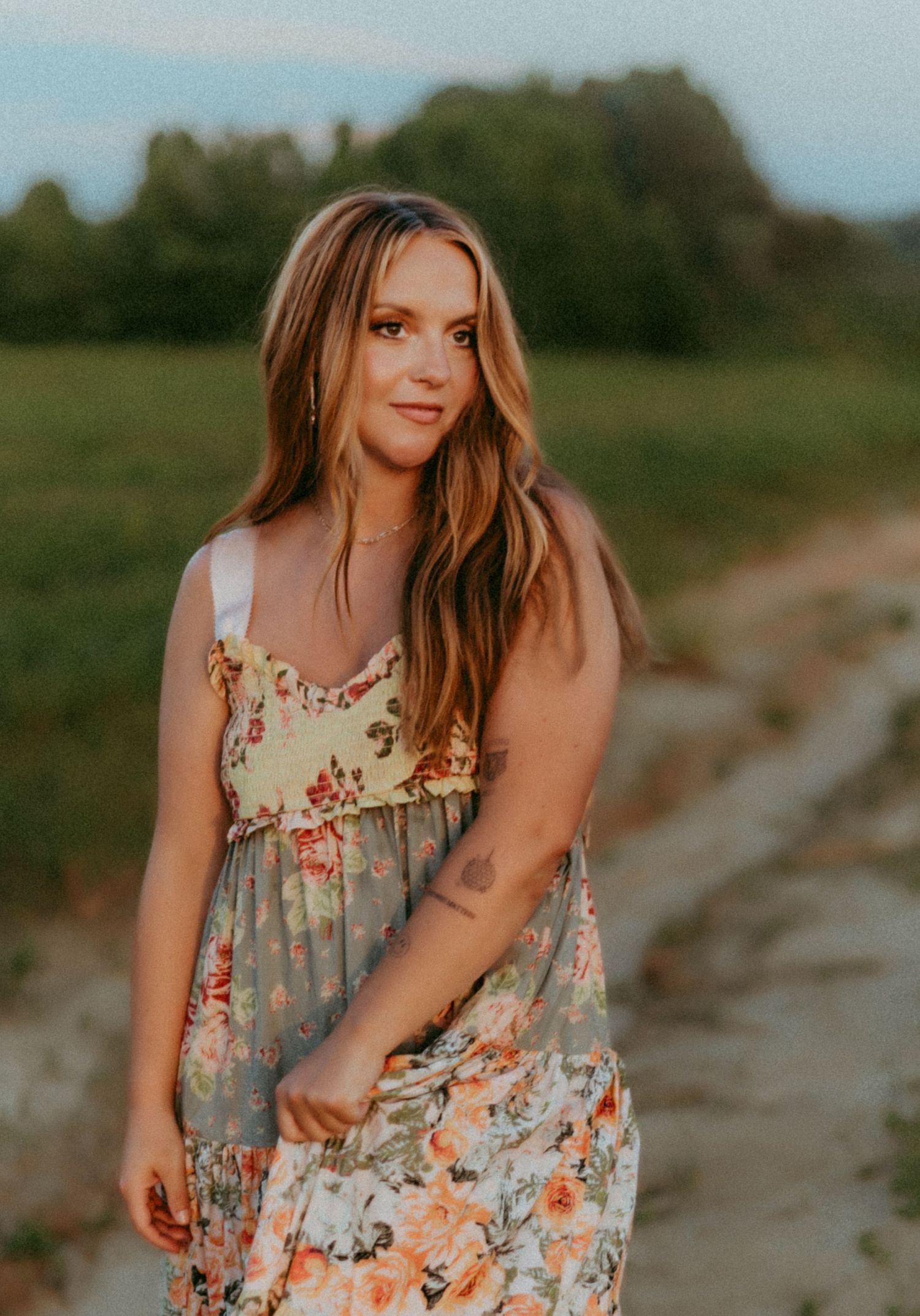 A person in a floral sundress poses outdoors at sunset in a natural setting with trees in the background.