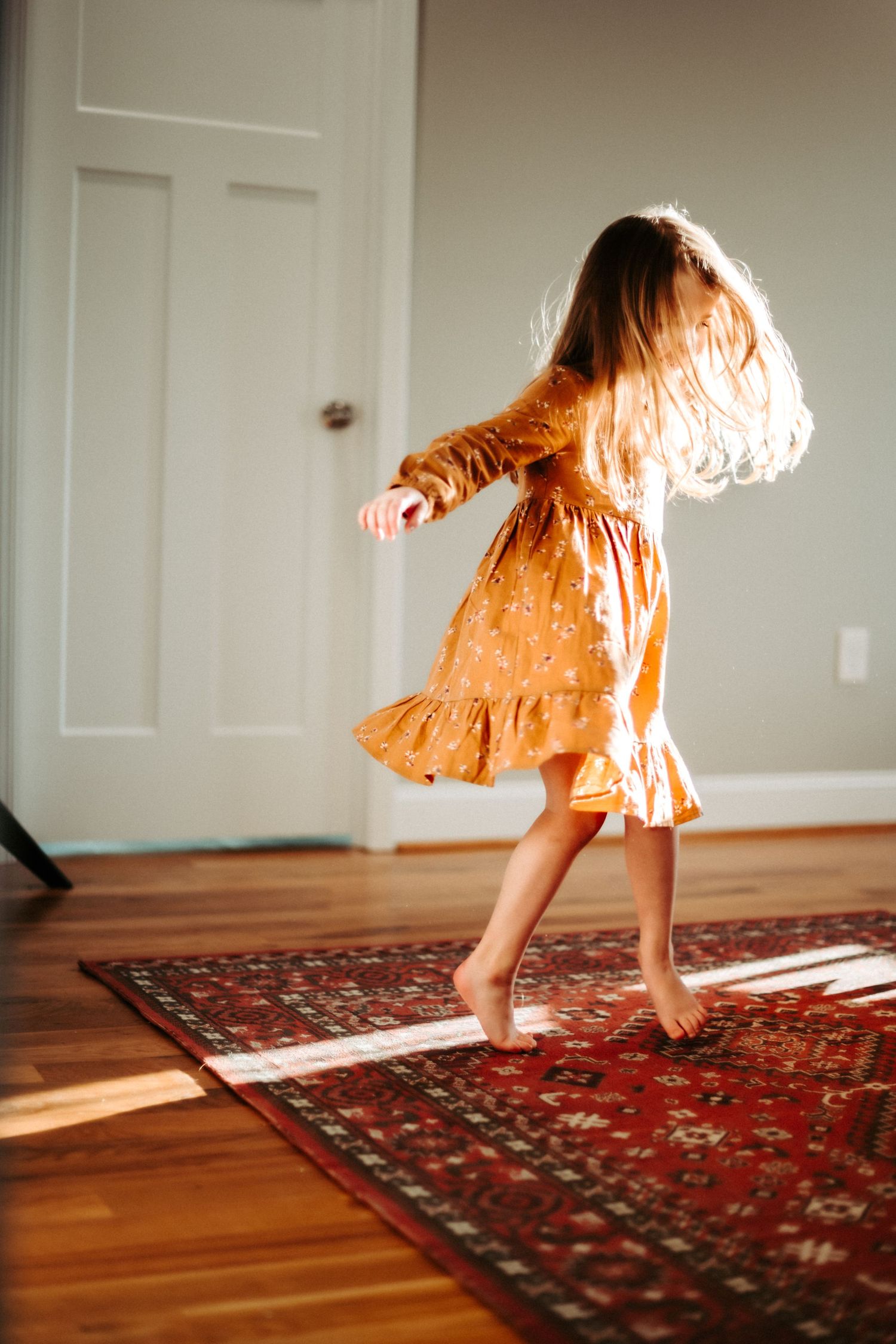 A barefoot figure in a flowing orange dress twirls and dances on hardwood floors over an ornate red rug in sunlit room.