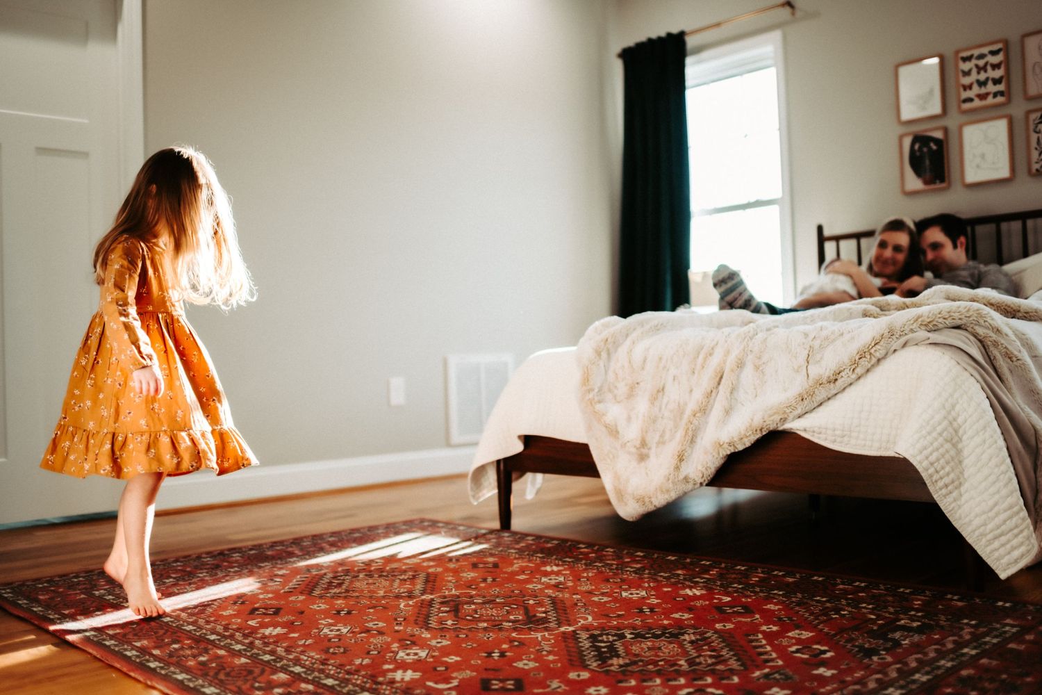 Bright sunlight streams through window onto child in orange dress standing on ornate red rug in bedroom.