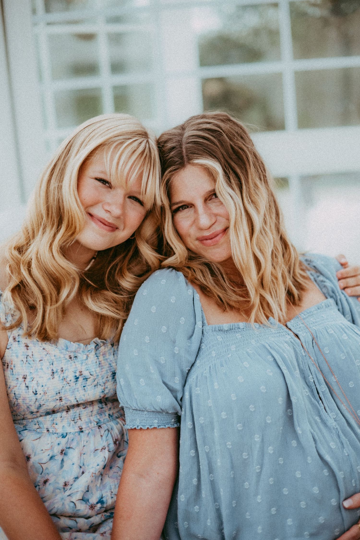 Two people with blonde wavy hair leaning together and smiling, wearing blue and floral summer dresses.