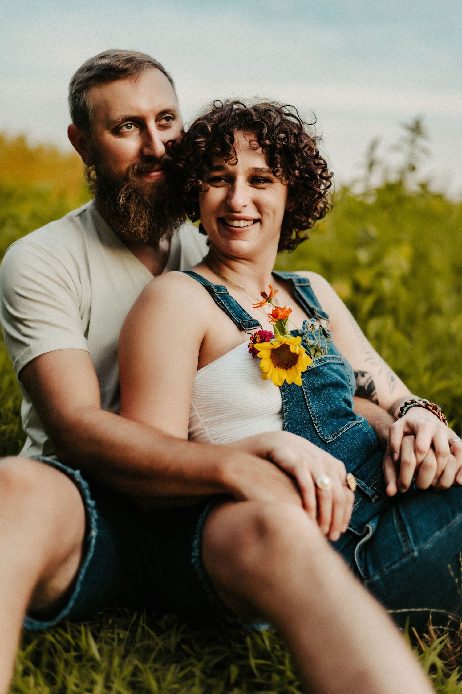 A couple sits together in a sunny field, the person in front wearing overalls with a sunflower accent.