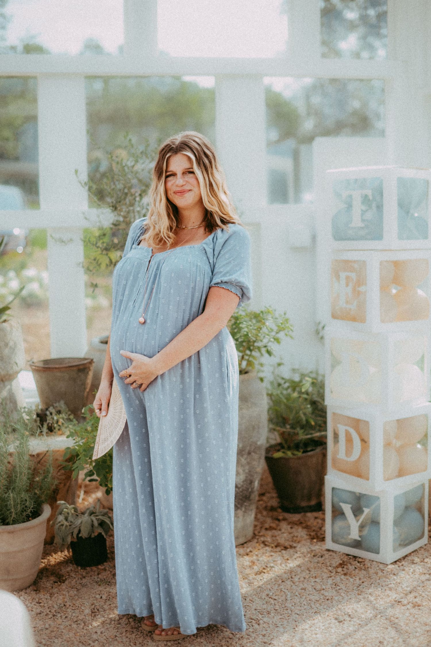 A person in a flowing grey maxi dress stands in a garden setting with decorative blocks visible in the background.