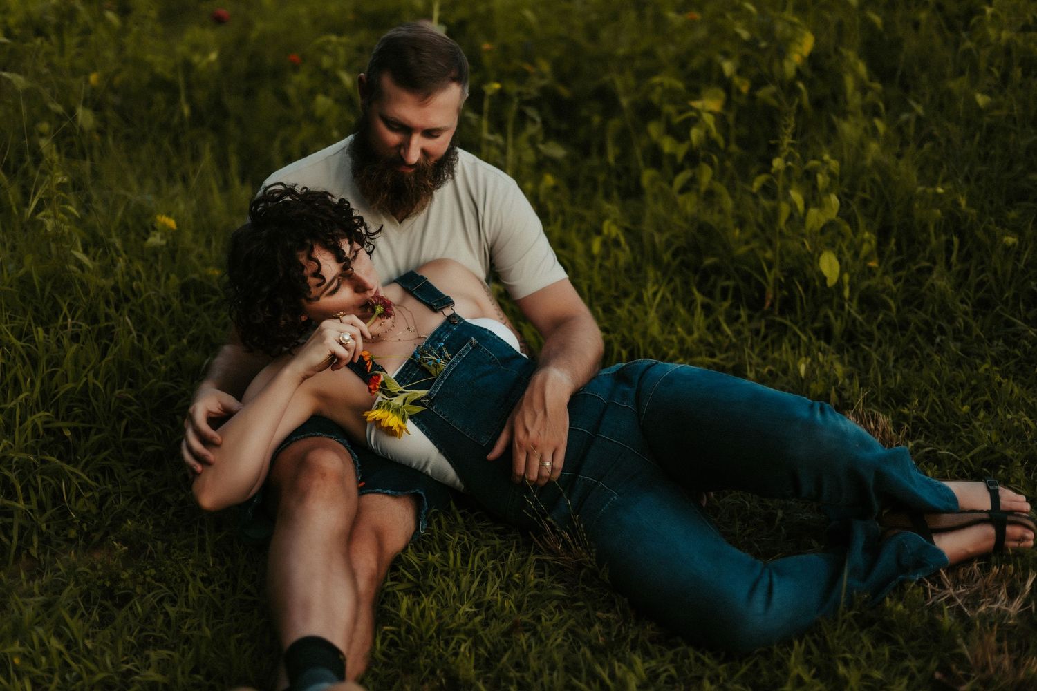 A couple rests together on grass during golden hour, with yellow wildflowers in a natural outdoor setting.
