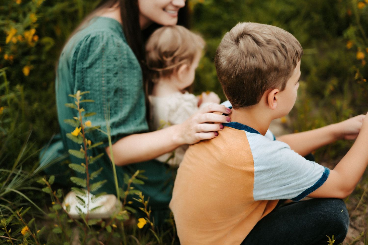 Family sits together in a field of yellow wildflowers on a warm sunny day wearing colorful casual clothing.