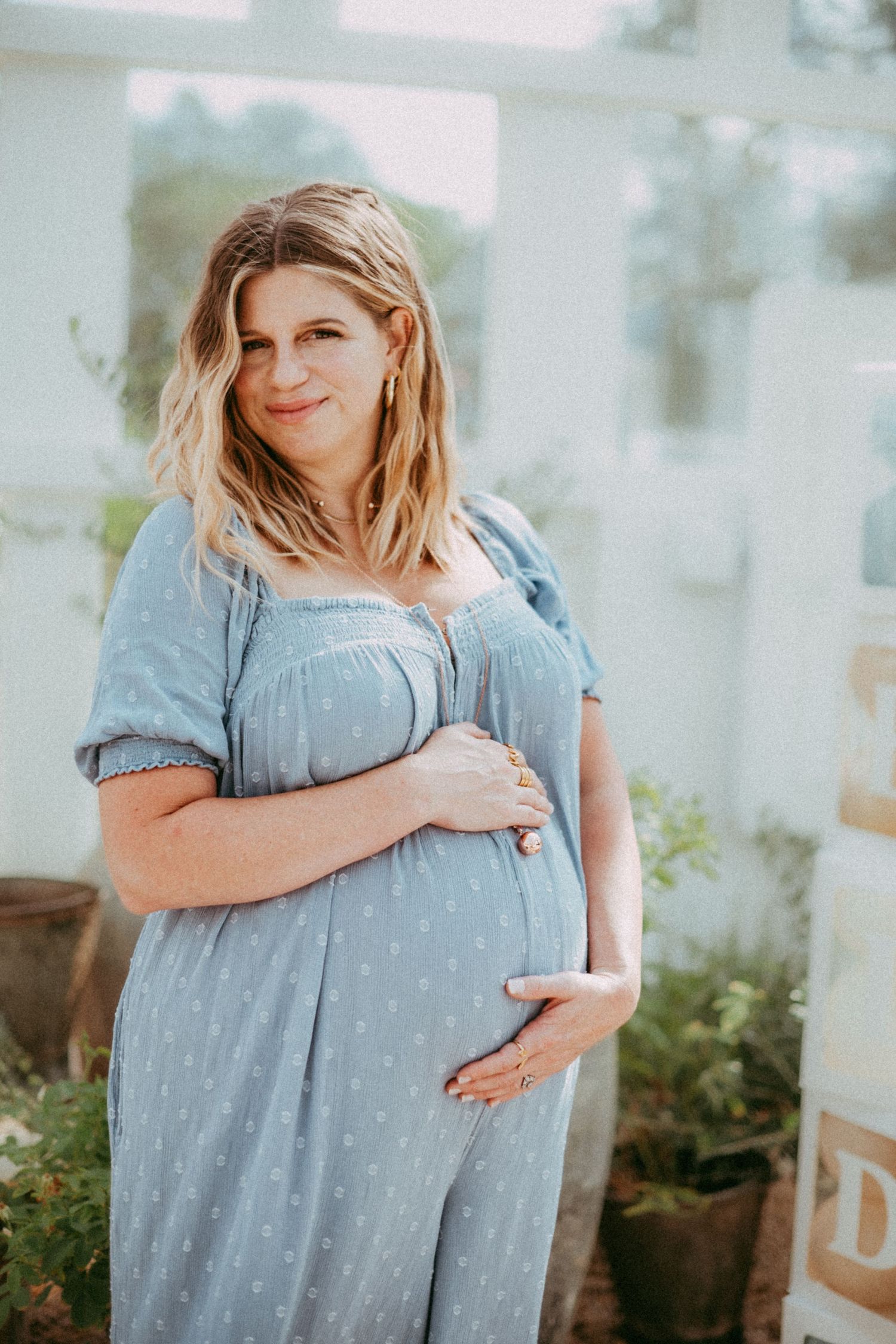 A pregnant woman in a blue polka dot dress poses outdoors while cradling her baby bump on a sunny day.
