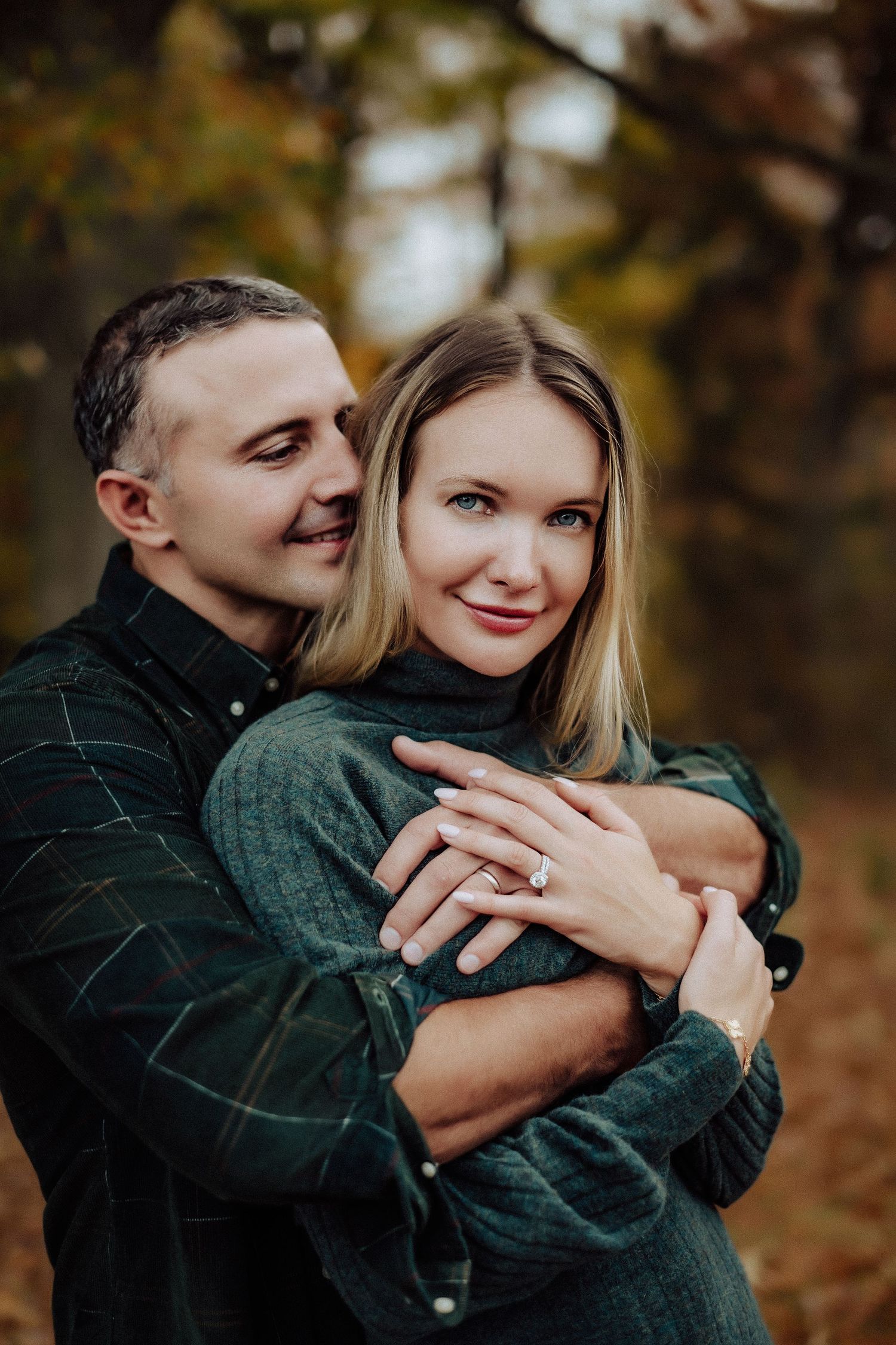 A romantic autumn engagement photo showing a couple embracing in cozy sweaters against a blurred forest background.