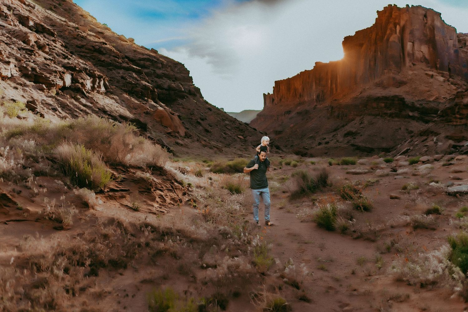 A hiker stands on a winding trail between towering red rock formations in a desert canyon landscape.