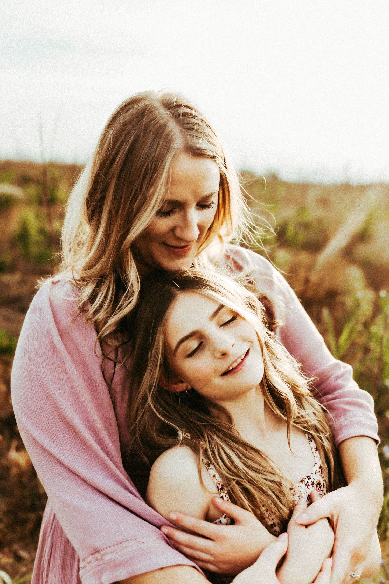 Two people in flowy dresses share a tender embrace while laughing together in a sunlit outdoor setting.