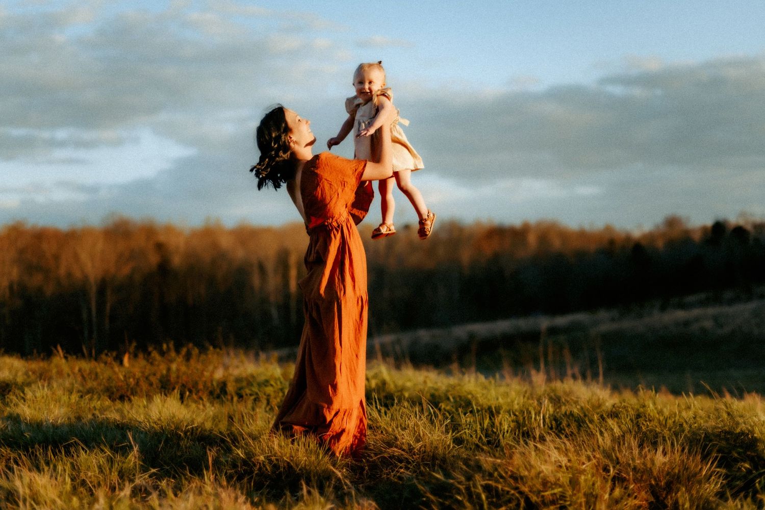 A person in a flowing orange dress lifts a small child into the golden sunset light on a grassy hilltop.