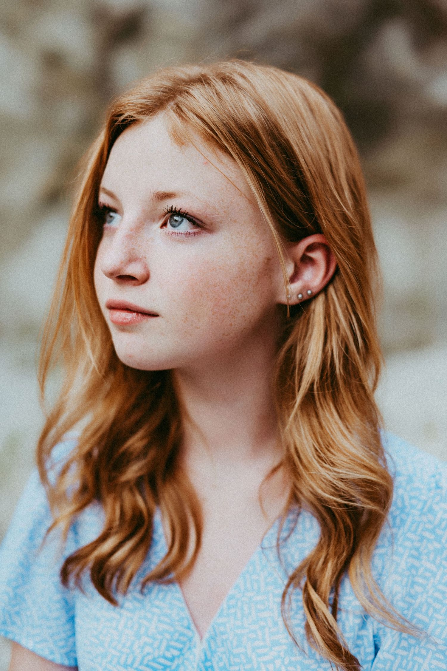 A young woman with strawberry blonde hair and freckles gazes off into the distance while wearing a light blue top.