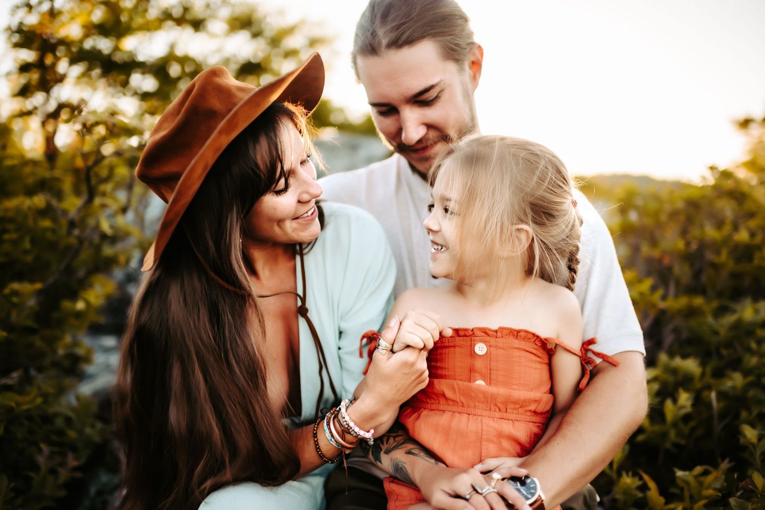Family enjoying a sunset moment together outdoors, with a brown hat and coral-colored dress against natural scenery.