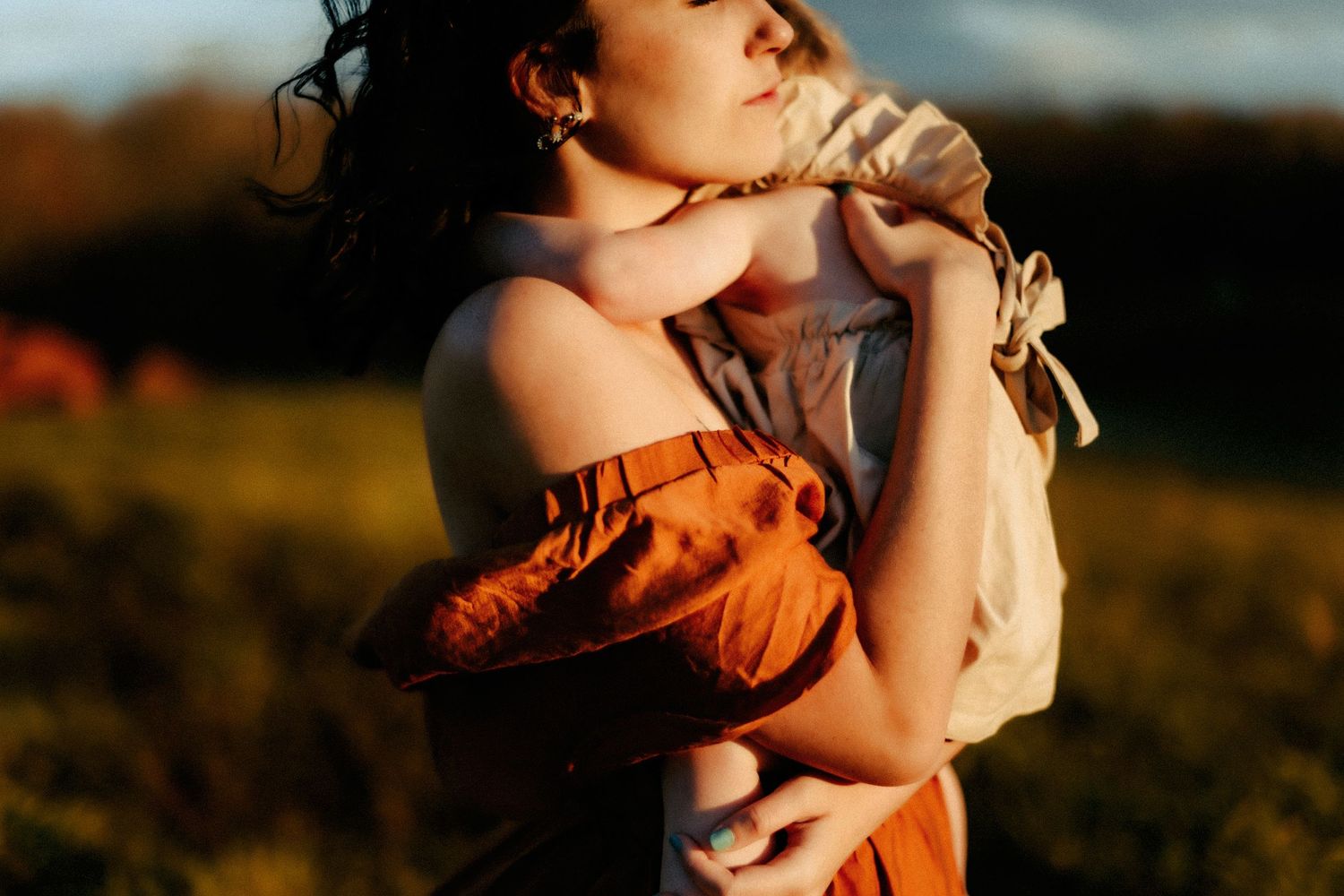 A tender moment captured at sunset as parent and baby embrace in warm golden light against a blurred outdoor backdrop.