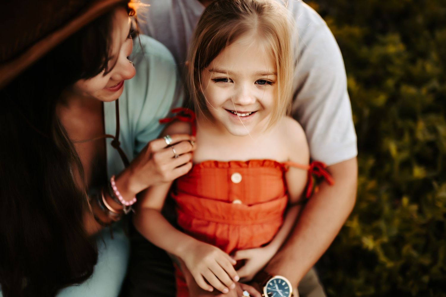 A child in an orange button-up dress smiles brightly while sitting outdoors in the summer.