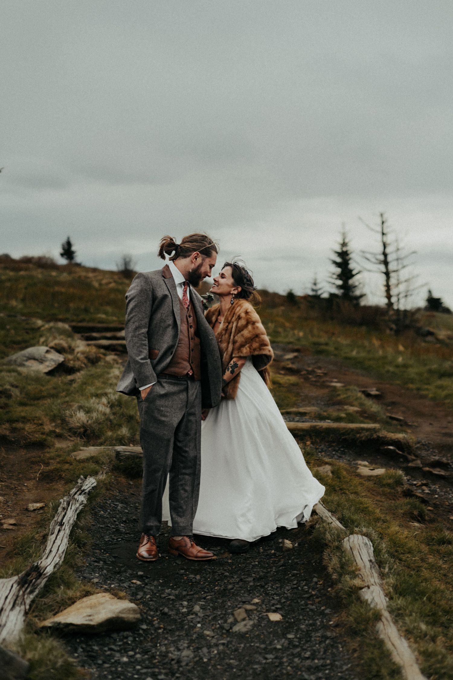 A couple embraces on a misty mountain trail wearing wedding attire and a plaid shawl against a moody grey sky.