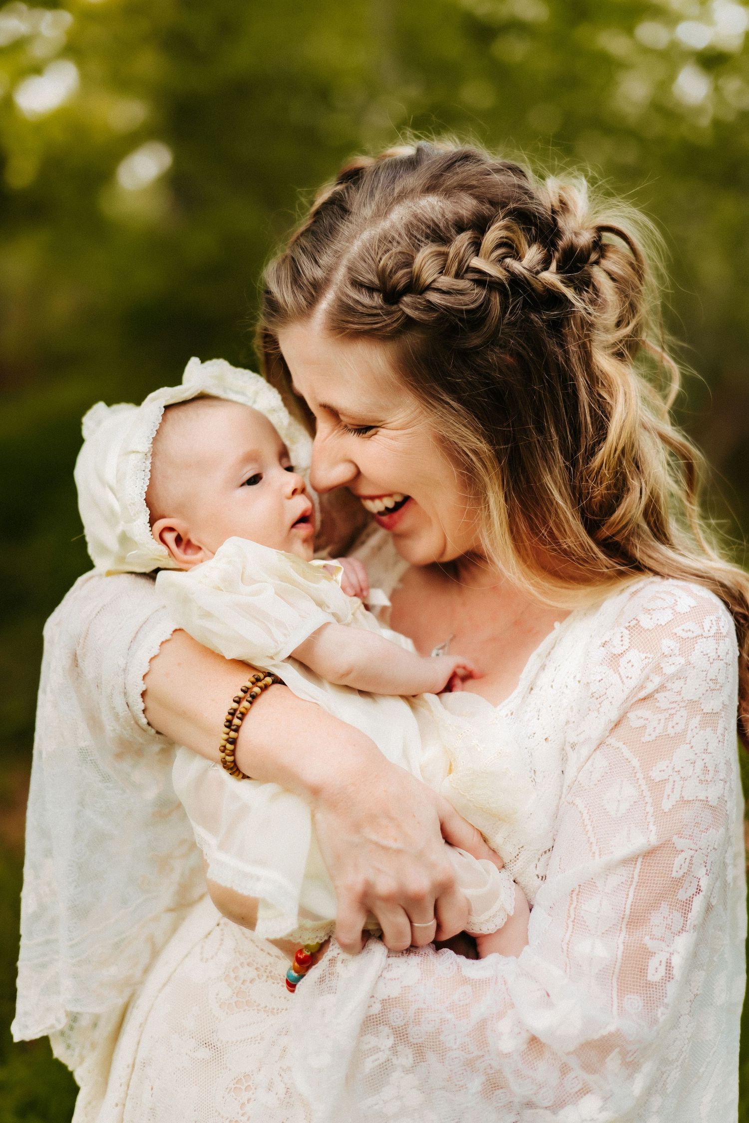 Joyful moment shared between a mother and baby in white lace dress with braided hair outdoors in nature.