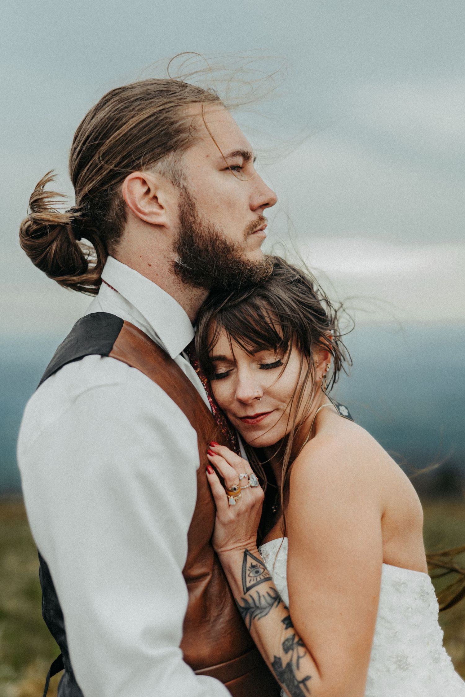 A newlywed couple shares an intimate moment outdoors with wind-swept hair against a cloudy mountain backdrop.