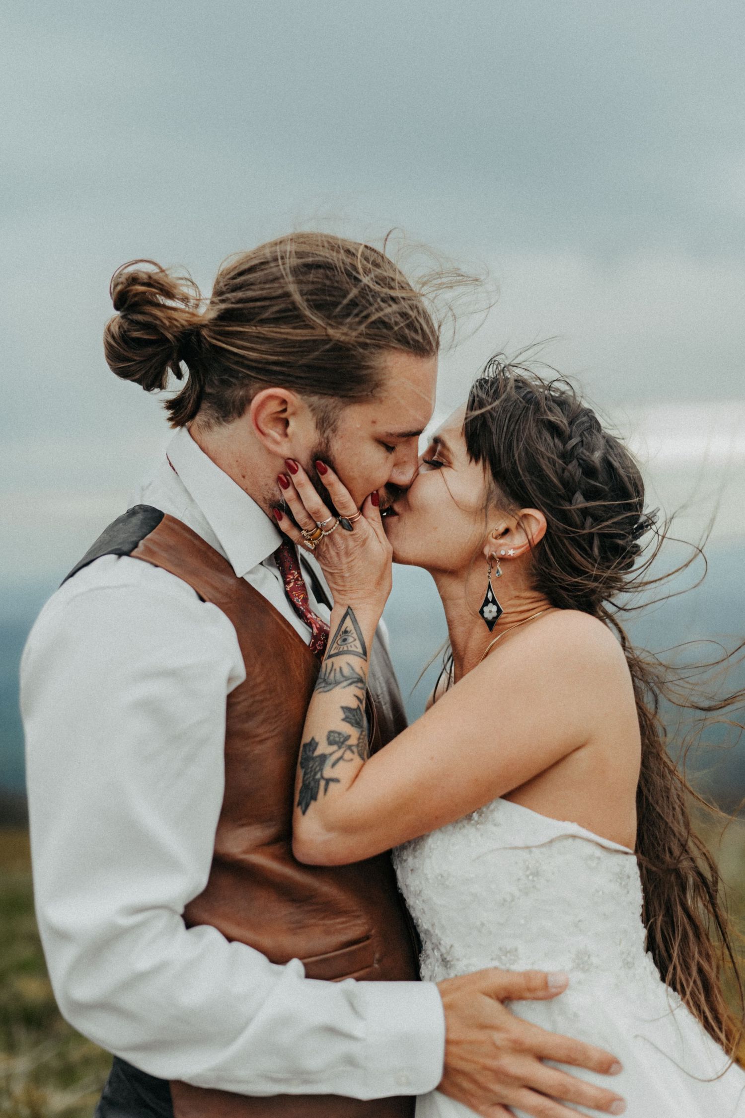 A couple shares a romantic kiss outdoors on their wedding day with wind-swept hair and cloudy skies in the background.