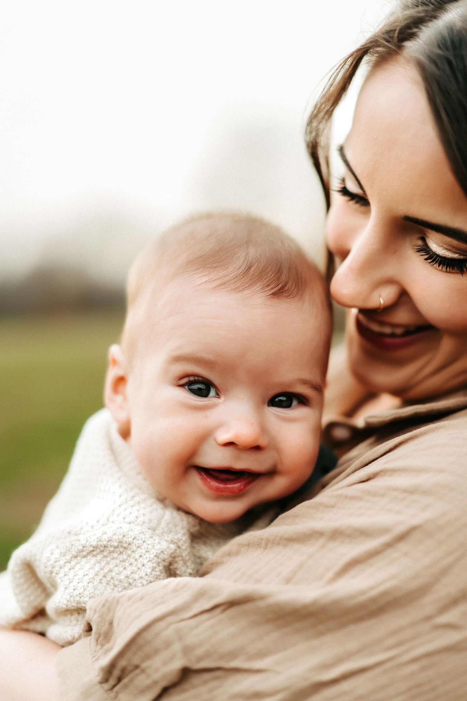 A smiling baby in a white sweater cuddles close with their mother in a tender outdoor portrait.