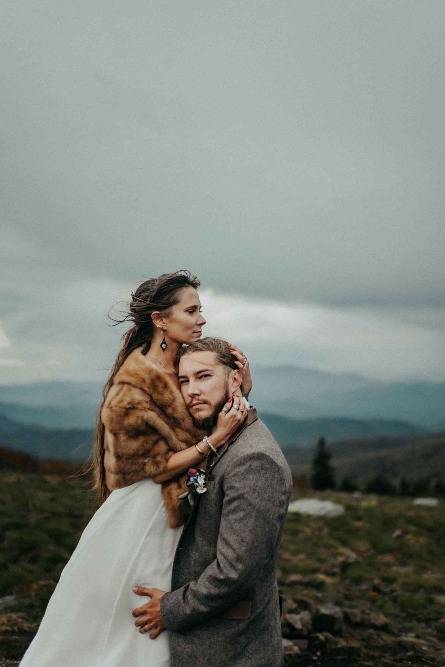 A couple embraces on a mountainside under moody gray skies, with the bride wearing a fur wrap over her wedding dress.