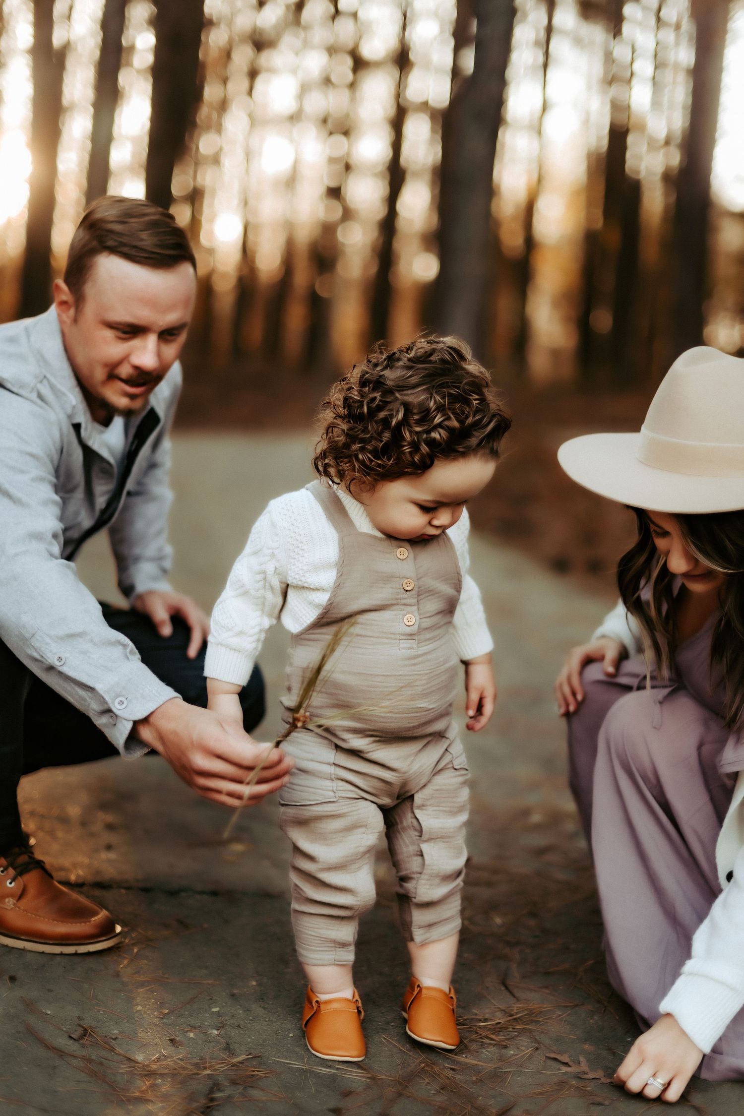 Toddler in beige outfit and brown shoes walks in forest at sunset while parents assist on either side.
