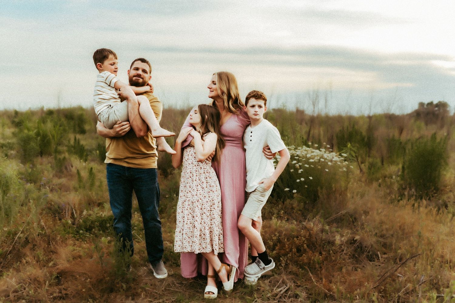 A family stands together in a natural field with tall grass during sunset in casual attire.