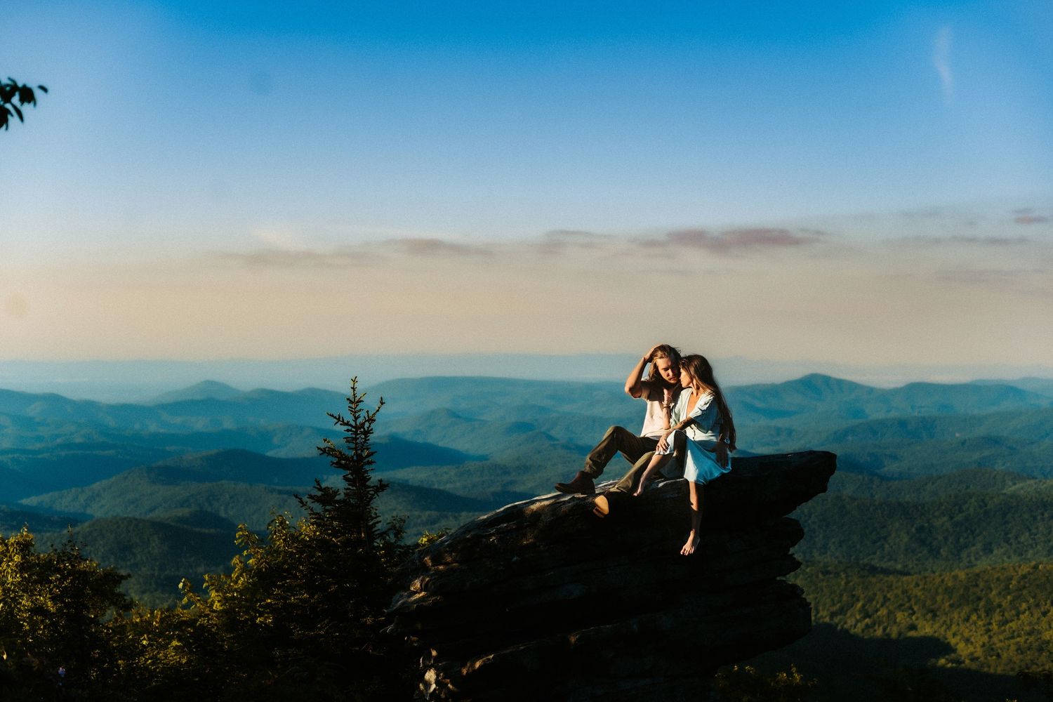 Two people sit together on a rocky overlook with mountain ranges and blue skies stretching to the horizon.