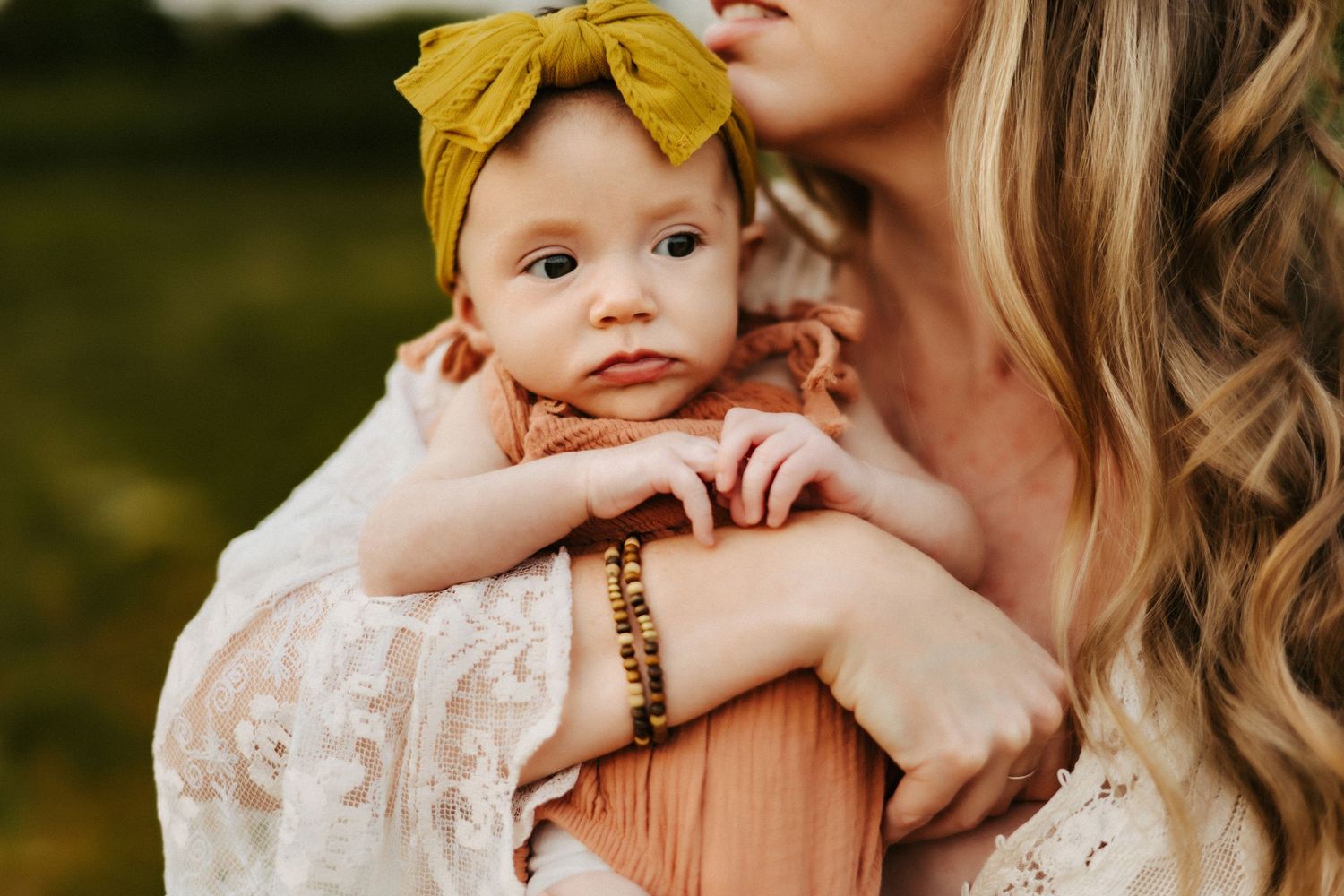 Infant wearing yellow headband is held close while wearing peachy-pink outfit against blurred outdoor background.