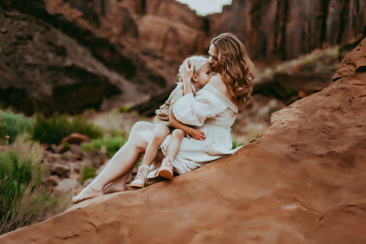 Two people in white outfits sit together on red rock formations at sunset in a desert canyon landscape.