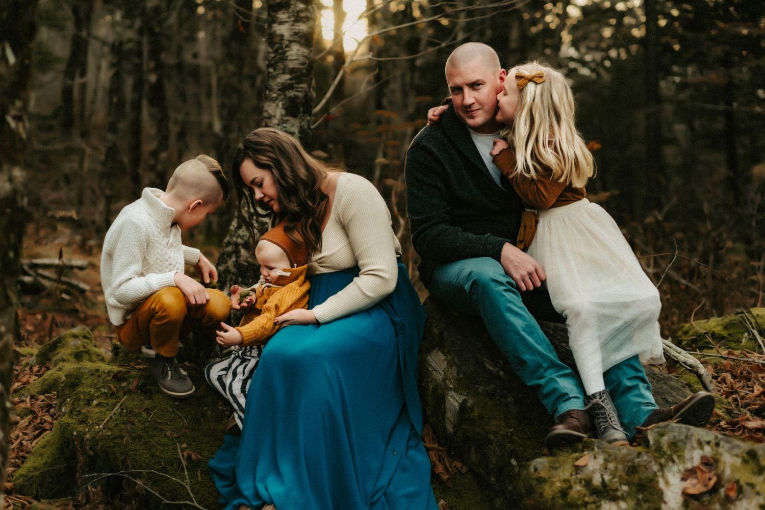 A family of four sits together on fallen logs in a misty autumn forest during golden hour.