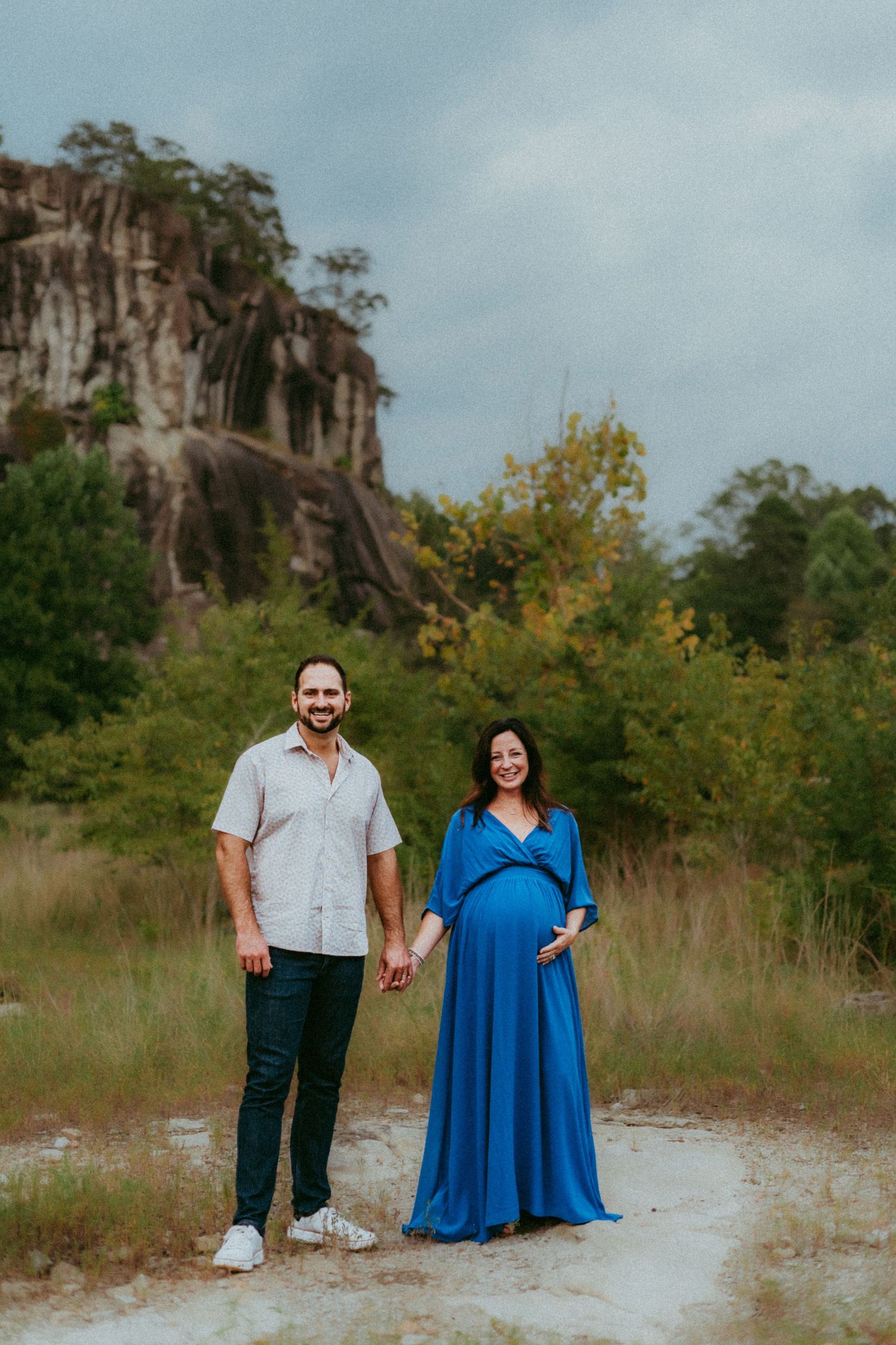 A couple stands holding hands on a dirt path with a scenic rocky cliff and autumn foliage in the background.
