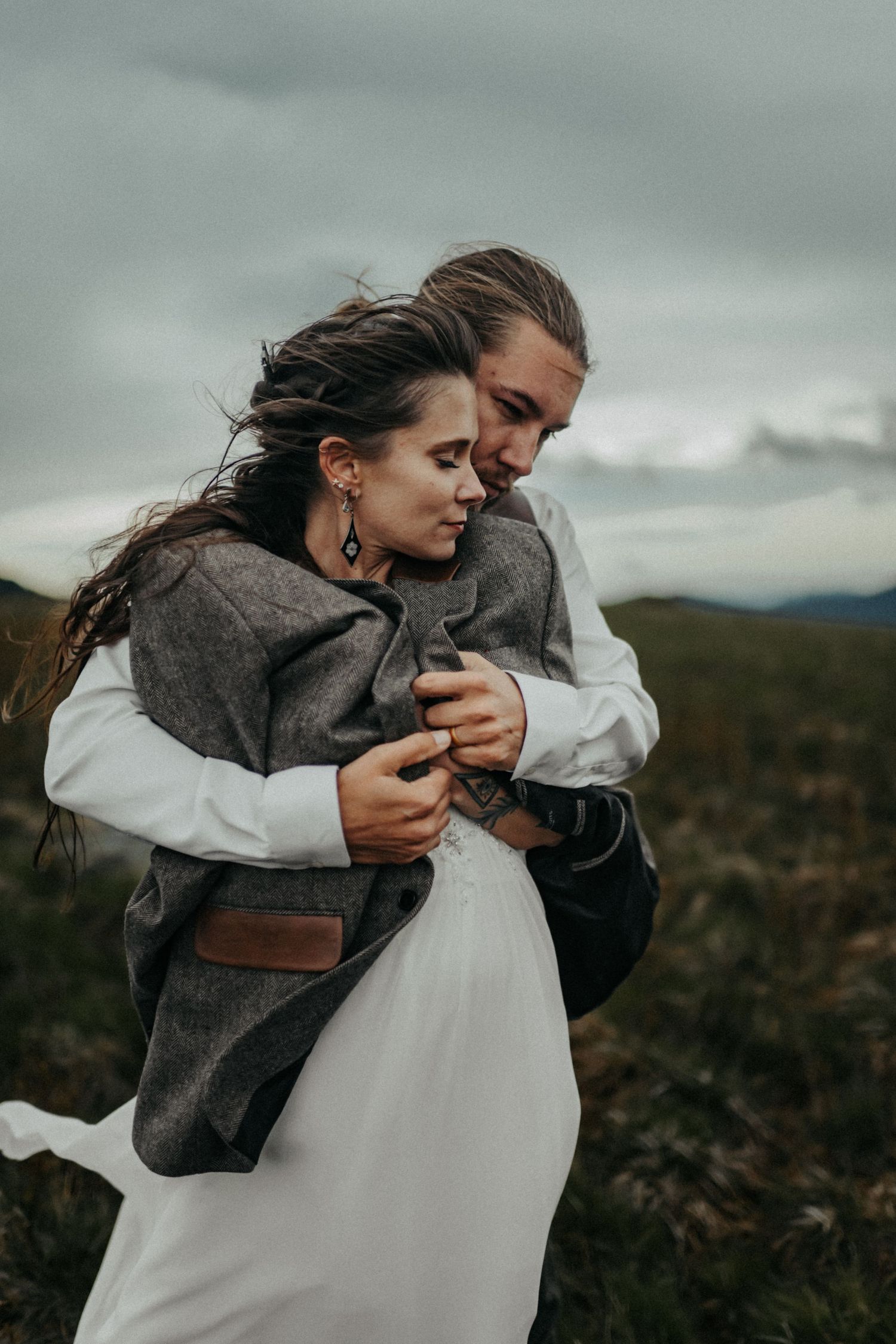 A romantic couple embraces outdoors on a cloudy day, with the woman wearing a white dress and gray shawl.