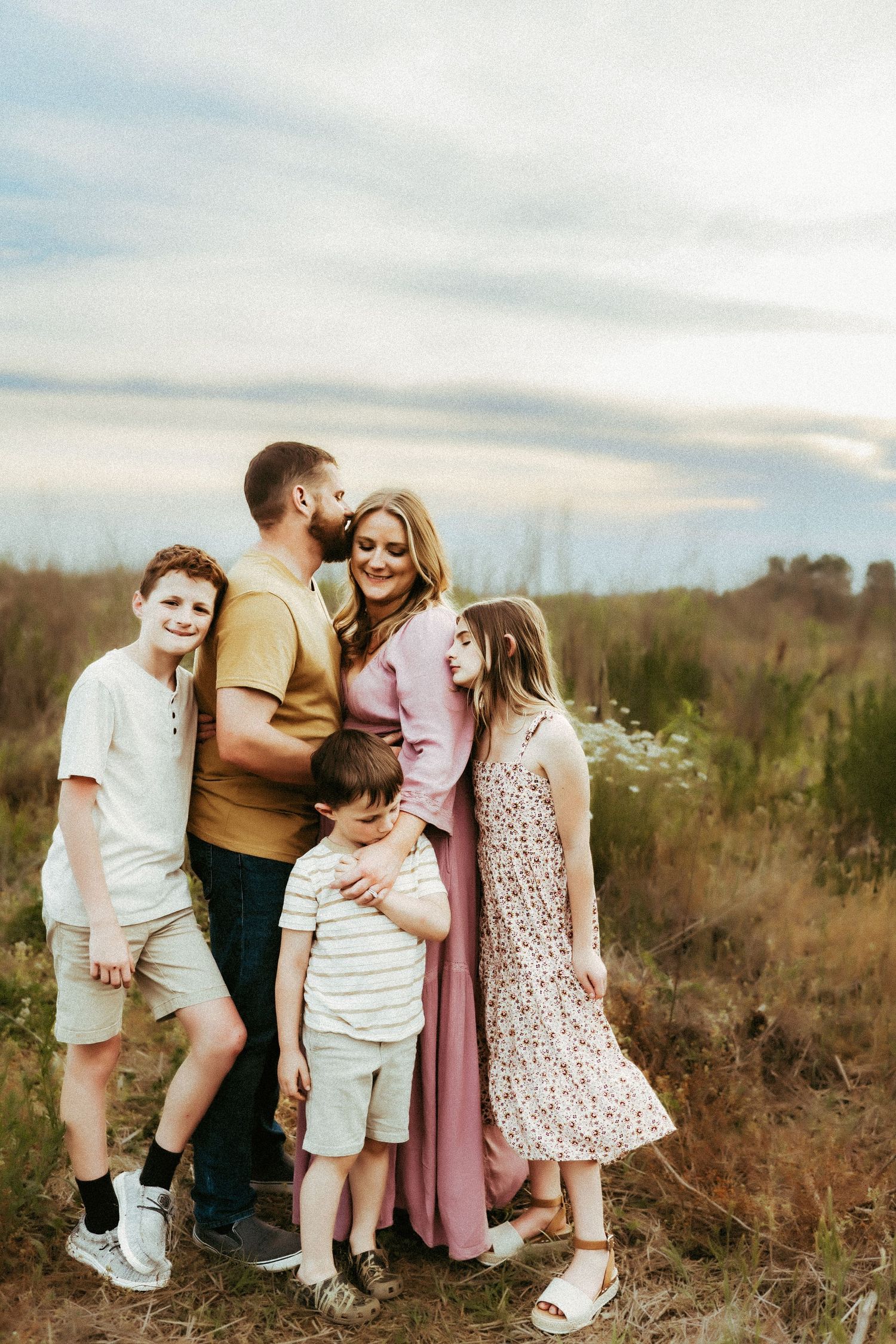 A family in casual clothing poses together outdoors at sunset in a natural field setting.